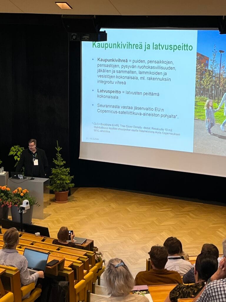 Puhuja pitää esitelmän konferenssissa auditoriossa, jossa on puiset istuimet. Hänen takanaan olevalla suurella kuvaruudulla näkyy suomenkielinen dia kaupunkien viherrakentamisesta, ja siinä on kuva ihmisistä, jotka kävelevät vihreiden ulkoleikkivälineiden lähellä. .