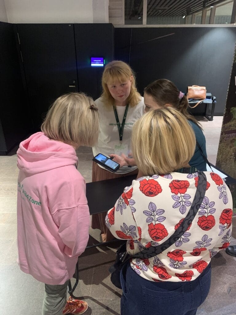 Neljä naista seisoo yhdessä sisätiloissa ja katselee älypuhelinta, jota yksi heistä pitää kädessään. He näyttävät keskustelevan. Yhdellä on yllään valkoinen paita ja lantioliina, kun taas muilla on värikkäät topit ja takit. .