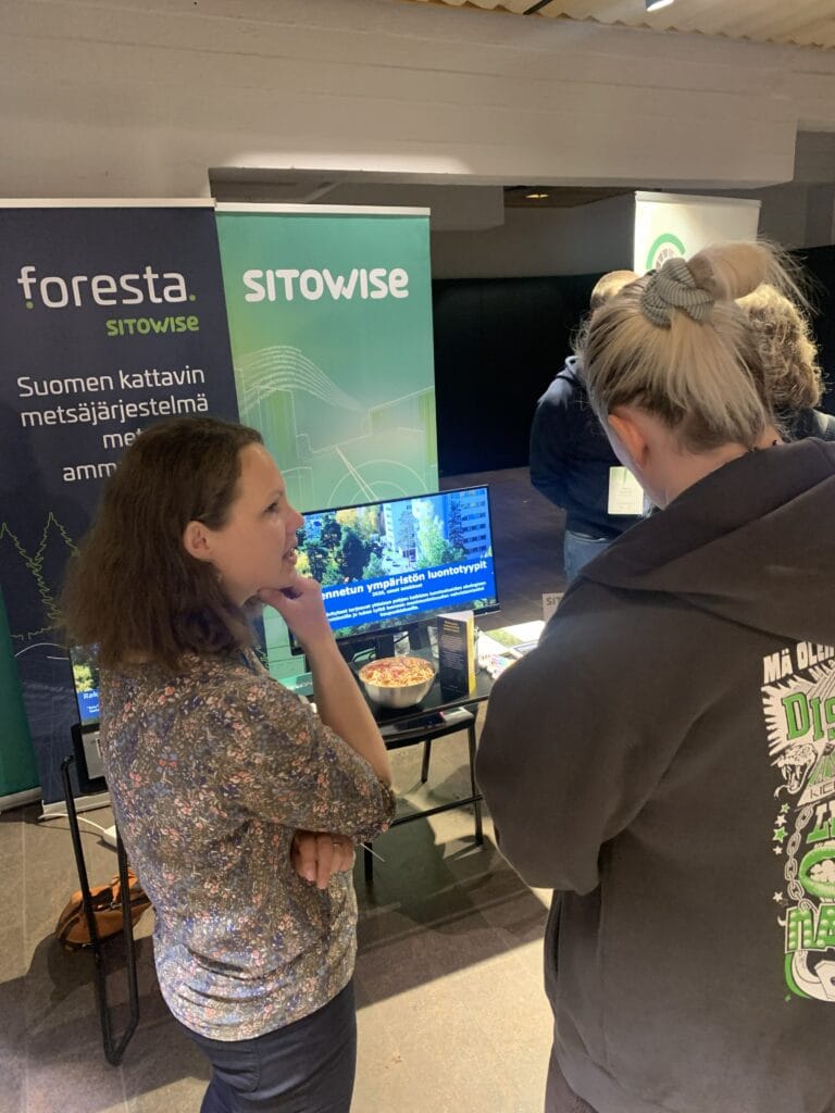 Kaksi naista seisoo ja keskustelee kojulla, jossa lukee "Foresta" ja "Sitowise". Näytöllä näkyy luontoon liittyvää sisältöä, ja pöydällä on tiedotusmateriaalia. Näyttää siltä, että kyseessä on sisätiloissa järjestettävä tapahtuma tai messutapahtuma. .