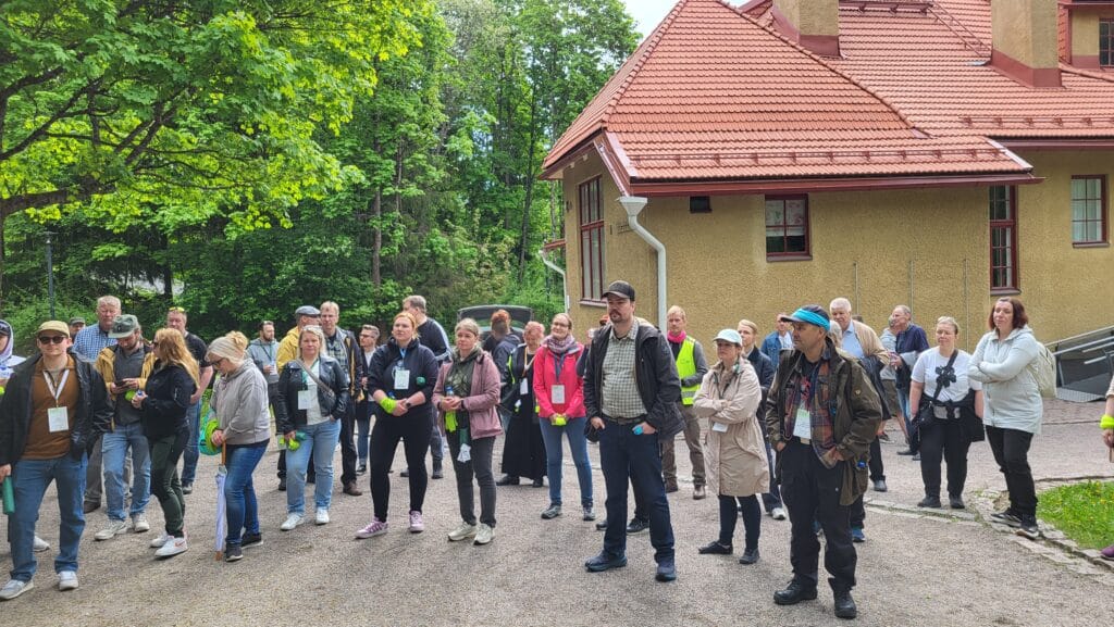 Joukko ihmisiä seisoo ulkona sorapolulla ja kuuntelee tarkkaavaisesti, taustalla on keltaisenruskea rakennus ja vihreitä puita. Monilla on nimilappuja ja vihreitä esineitä kädessään, mikä viittaa järjestettyyn tapahtumaan tai kierrokseen. .