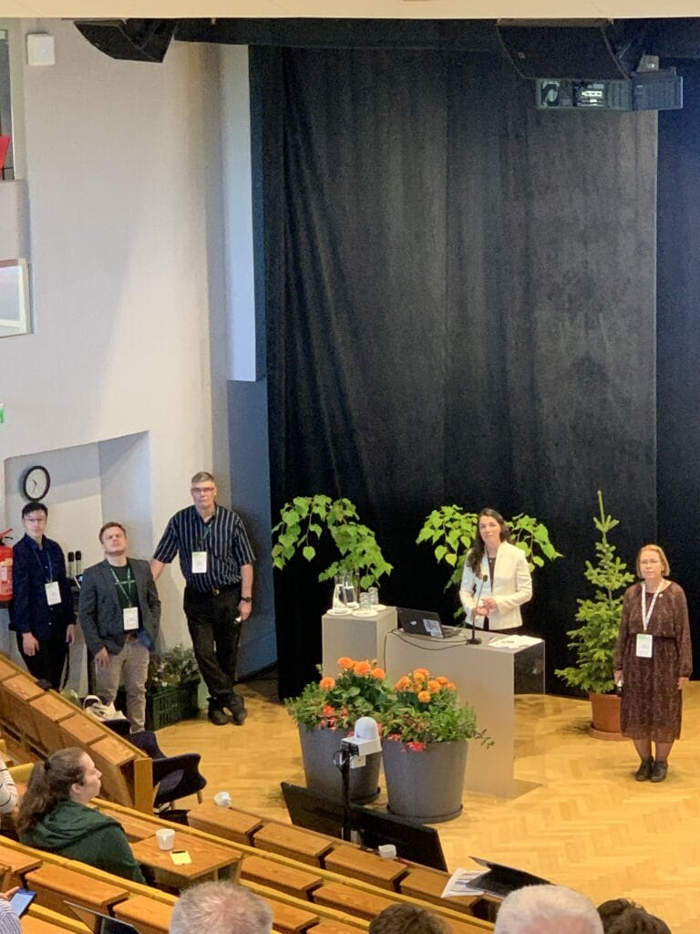Nainen seisoo korokkeella pitämässä esitelmää luentosalissa, ja viisi ihmistä seisoo lähellä. Ruukkukasvit ja kukat koristavat lavaa, ja yleisö istuu puuriveissä. .