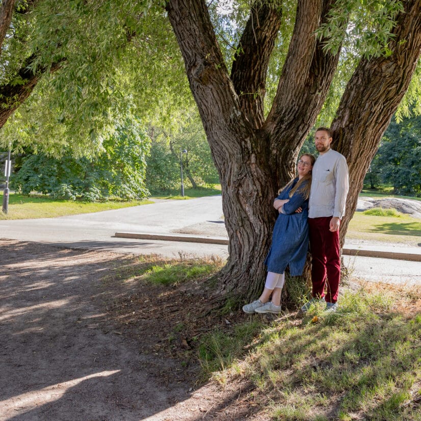 Pari seisoo tiiviisti vierekkäin suurten puiden varjossa aurinkoisessa puistossa, jonka taustalla näkyy vihreää nurmikkoa, kävelyreittiä ja kaukana olevia puita. .