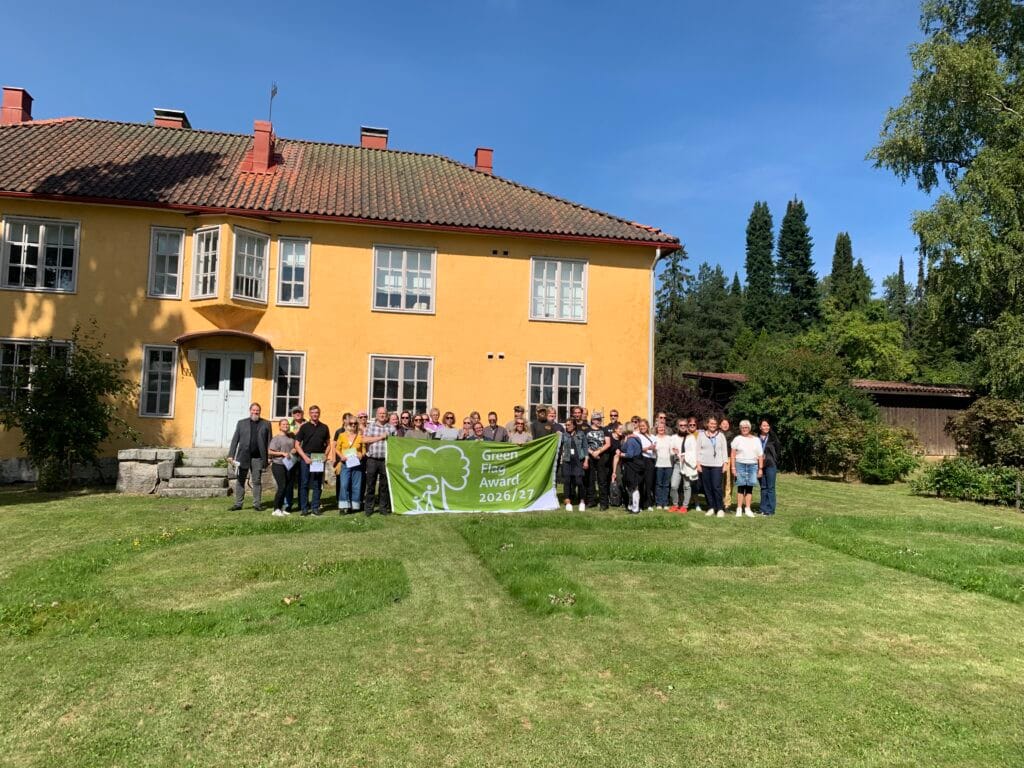 Ryhmä ihmisiä poseeraa aurinkoisena päivänä suuren keltaisen talon edessä pitäen kädessään vihreää Green Flag Award 2026/27 -banderollia. Nurmikko on siististi leikattu, ja rakennusta ympäröivät puut. .