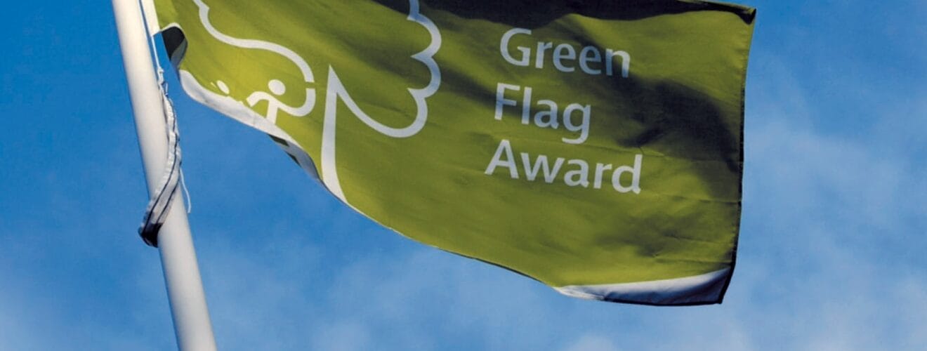 Vihreä lippu, jossa on valkoinen puun ääriviiva ja teksti ”Green Flag Award”, liehuu korkealla lipputangolla sinistä taivasta ja muutamia pilviä vasten. .