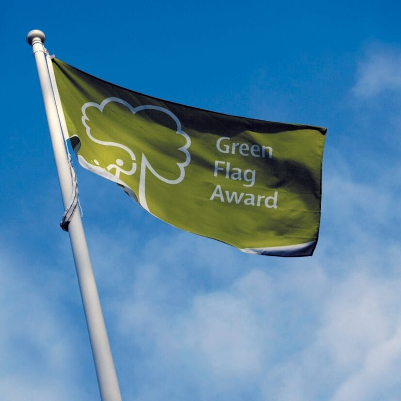 Vihreä lippu, jossa on valkoinen puun ääriviiva ja teksti ”Green Flag Award”, liehuu korkealla lipputangolla sinistä taivasta ja muutamia pilviä vasten. .