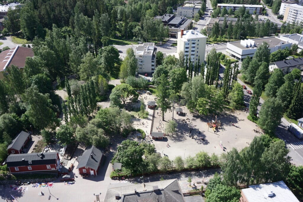 Ilmakuva puistosta, jossa on puita, leikkikentän välineitä ja ihmisiä. Puistoa ympäröivät asuinrakennukset, tiet sekä useita punavalkoisia taloja. Alue on vehreä. .