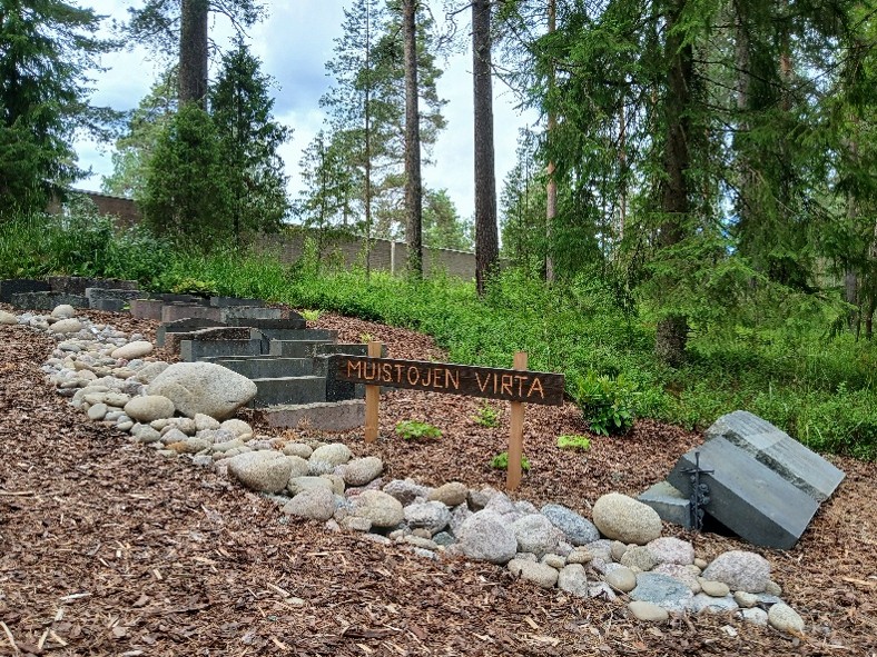 Kivillä reunustettu polku ja "Muistojen virta" -kyltti metsäisellä hautausmaan muistolehtoalueella.